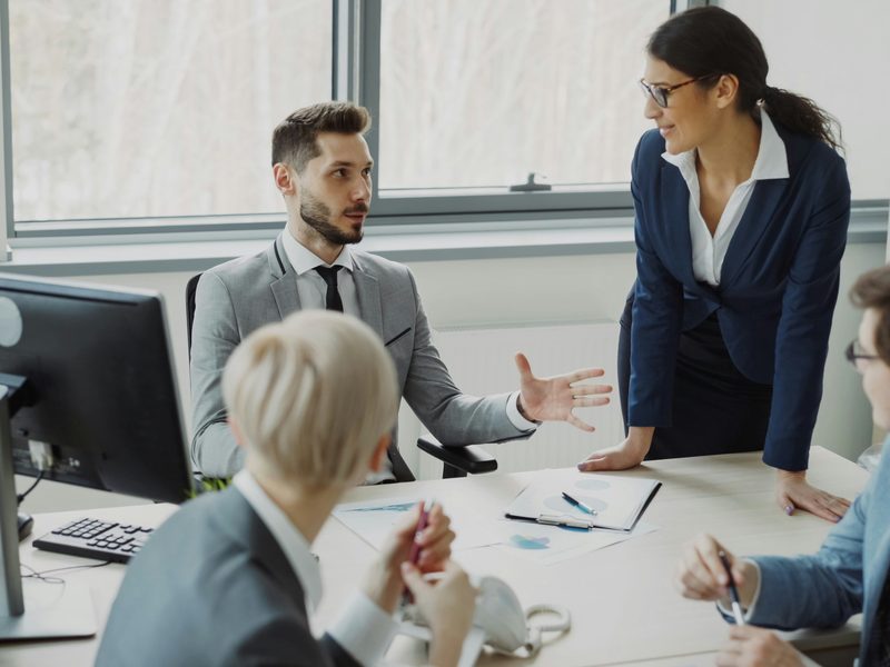Cuatro profesionales conversan alrededor de una mesa de reunión en una oficina luminosa; uno gesticula mientras explica y sobre la mesa hay gráficos impresos. Foto: Vitaly Gariev / Unsplash