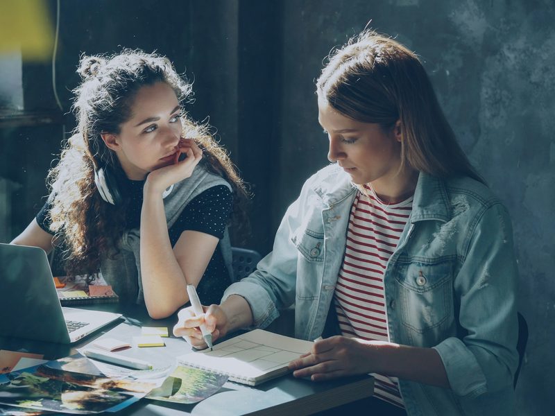 Dos emprendedoras trabajan juntas en un escritorio con una laptop, bocetos y notas adhesivas; una toma apuntes mientras la otra escucha. Foto: Vitaly Gariev / Unsplash