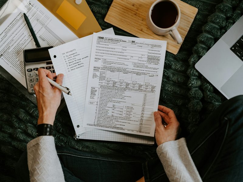 Vista desde arriba de una persona revisando papeles de finanzas personales, con una calculadora, una lista de documentos, un café y una laptop. Foto: Kelly Sikkema / Unsplash