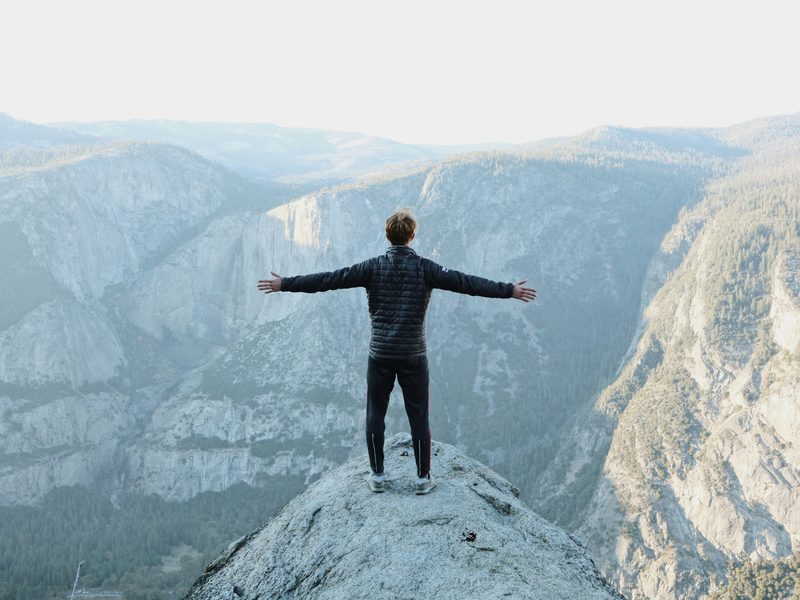 Una persona de espaldas, parada sobre una roca en la cima, abre los brazos frente a un valle de montañas al amanecer. Foto: Jason Hogan / Unsplash