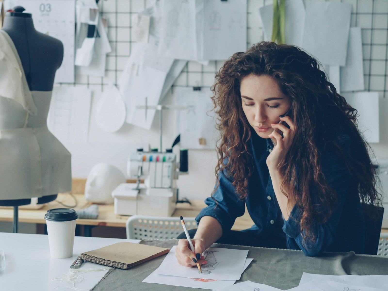 Una emprendedora trabaja en su taller: boceta a mano sobre la mesa mientras habla por teléfono, con un maniquí de costura y patrones colgados detrás. Foto: Vitaly Gariev / Unsplash